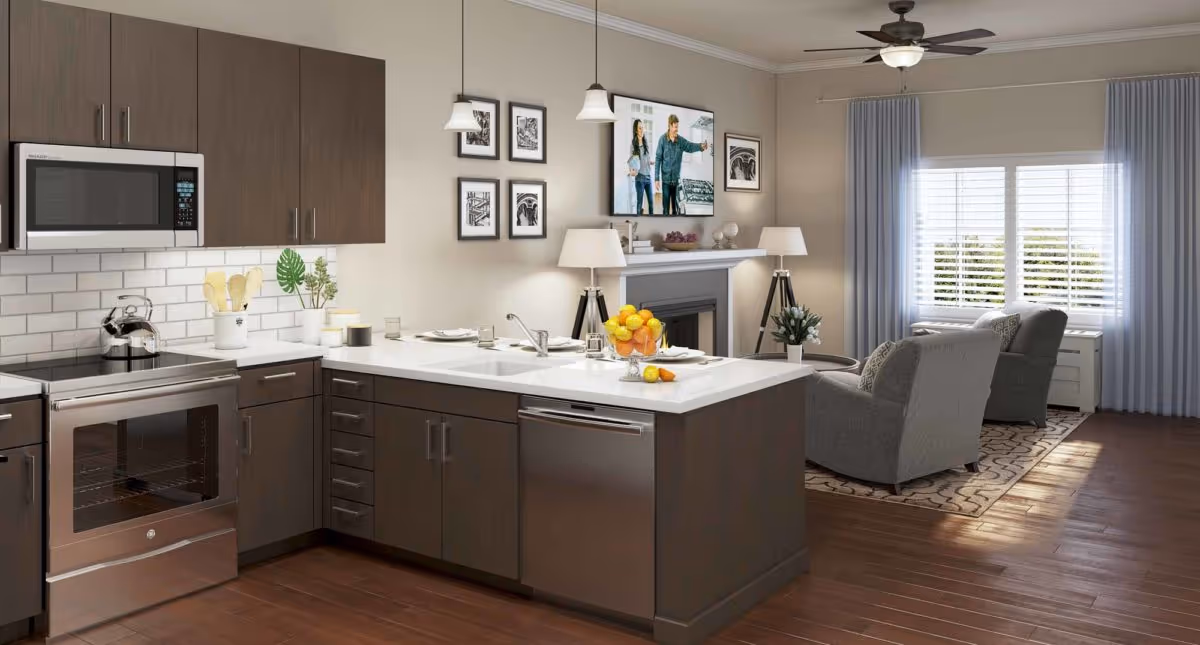 Modern open-concept kitchen and living room area with dark wood cabinets, stainless steel appliances including a microwave and oven, a white countertop island with a sink and a bowl of fruit, two gray armchairs on a patterned rug, a fireplace with a TV mounted above it, and large windows with light blue curtains letting in natural light.