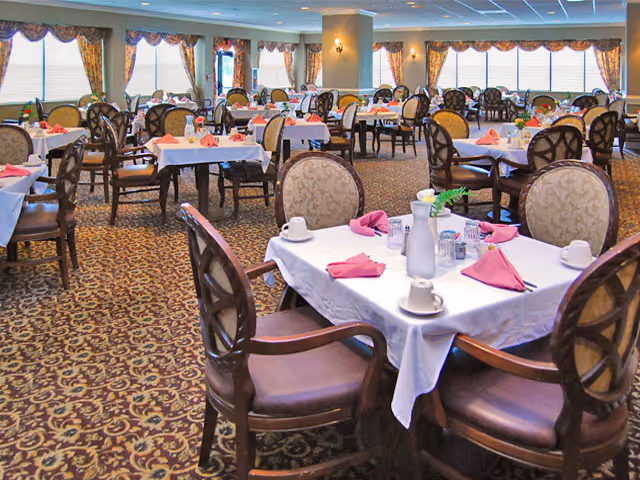 A spacious dining room with multiple round tables covered with white tablecloths, each set with pink folded napkins, cups, and small centerpieces. The room has patterned carpet flooring, large windows with curtains, and elegant chairs surrounding the tables.