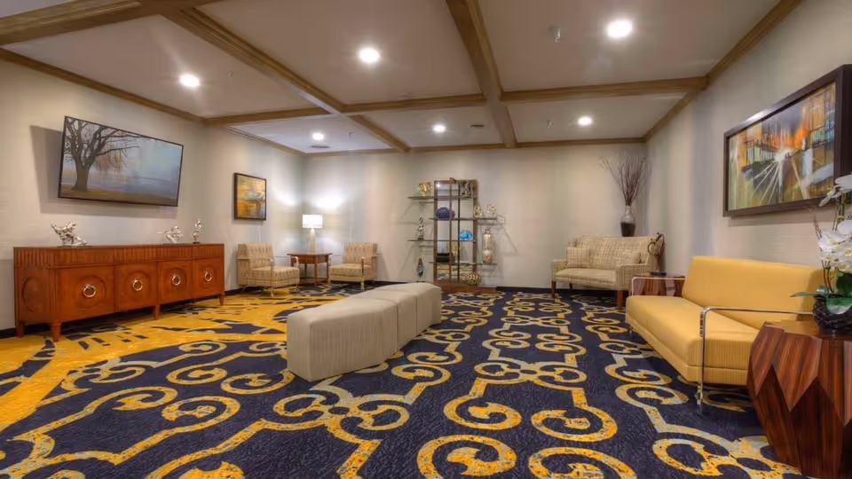 A senior living facility lounge with patterned carpet, sofas and chairs, a long ottoman, wooden credenza and wall art under a coffered ceiling.