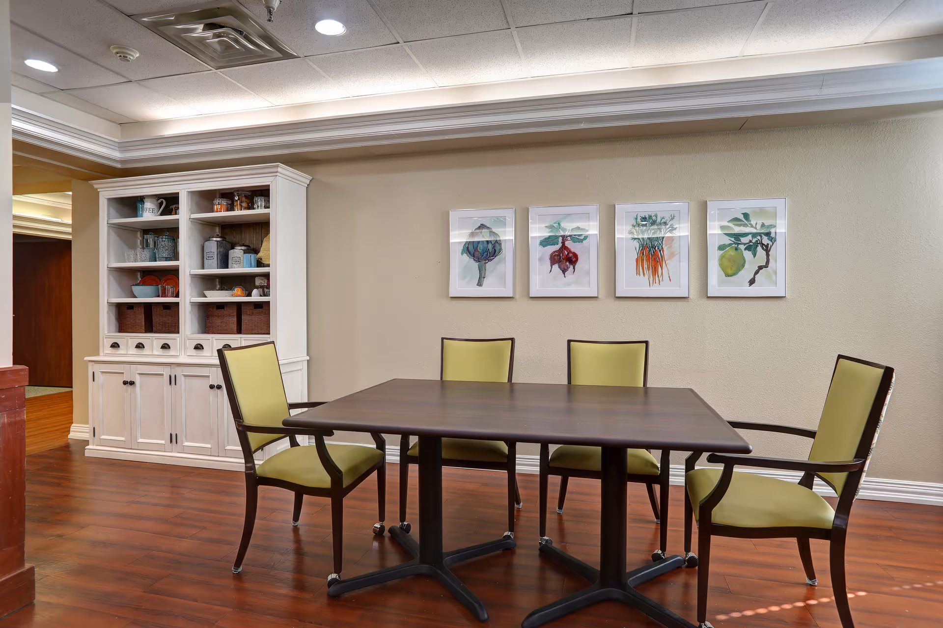Dining area with a rectangular table surrounded by green-upholstered chairs, a white cabinet on the left, and four framed botanical prints on the wall.