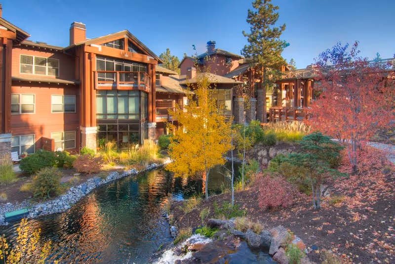 Exterior view of a senior living facility with rustic wooden architecture surrounded by trees with autumn foliage and a small pond with rocks and plants.