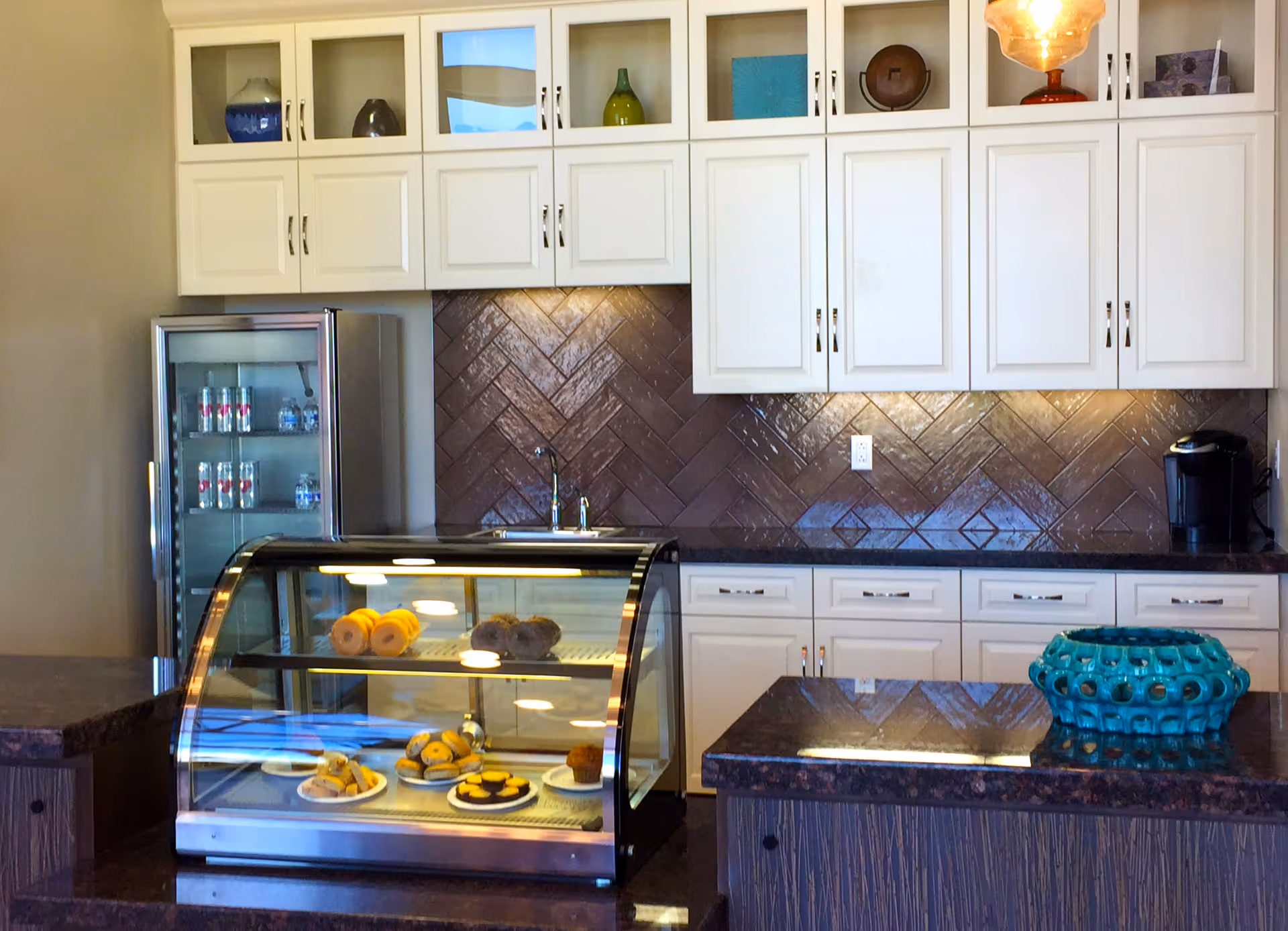 Interior view of a kitchen area with white cabinets, a glass-front refrigerator stocked with bottled water and canned drinks, a display case with various pastries, and a countertop with a decorative blue ceramic bowl.