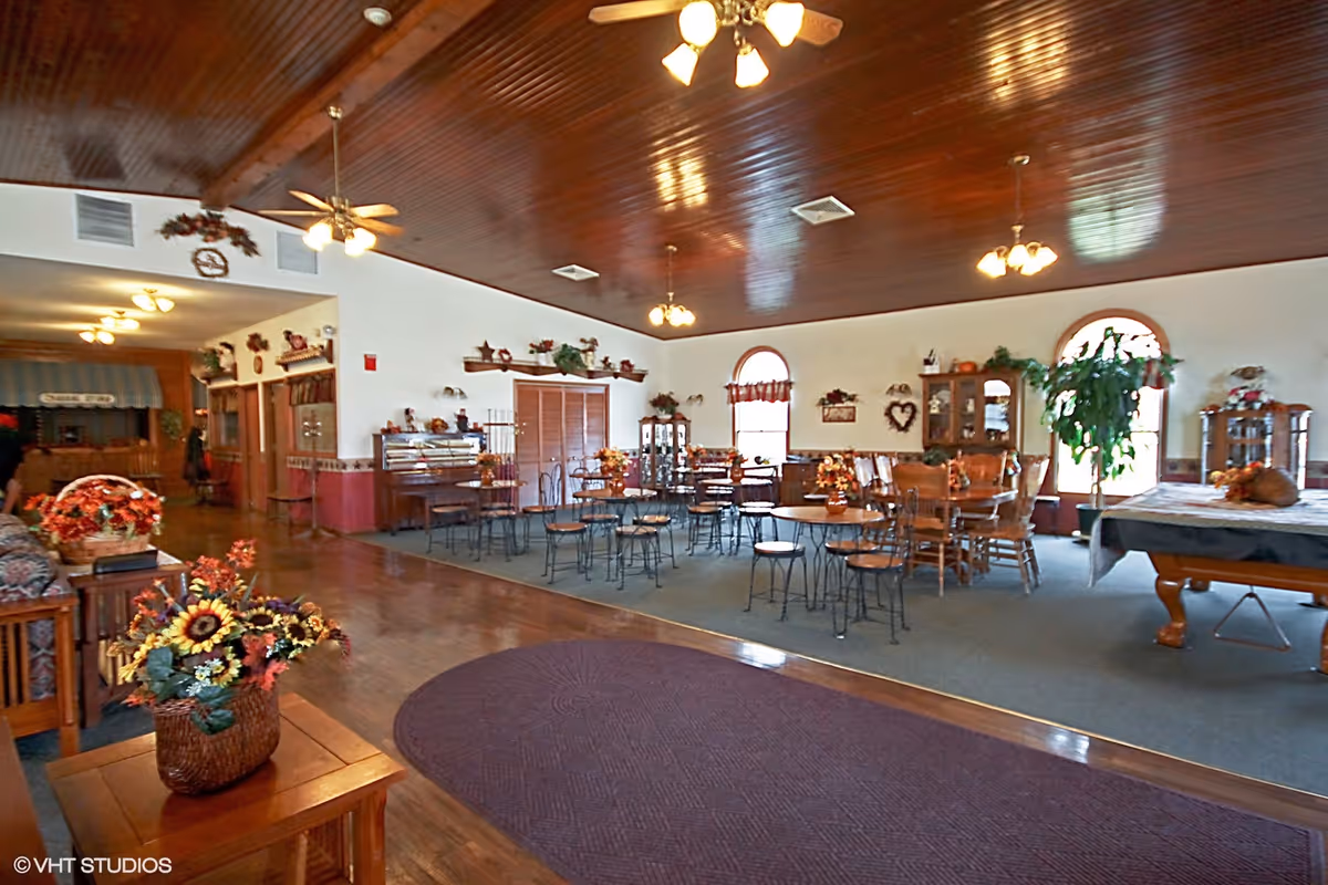 Spacious common area with multiple round tables and chairs, decorated with autumn-themed floral arrangements. The room features a wooden ceiling with ceiling fans and light fixtures, large arched windows with curtains, and various wooden cabinets and shelves with decorative items. There is a pool table on the right side and a carpeted area in the center.