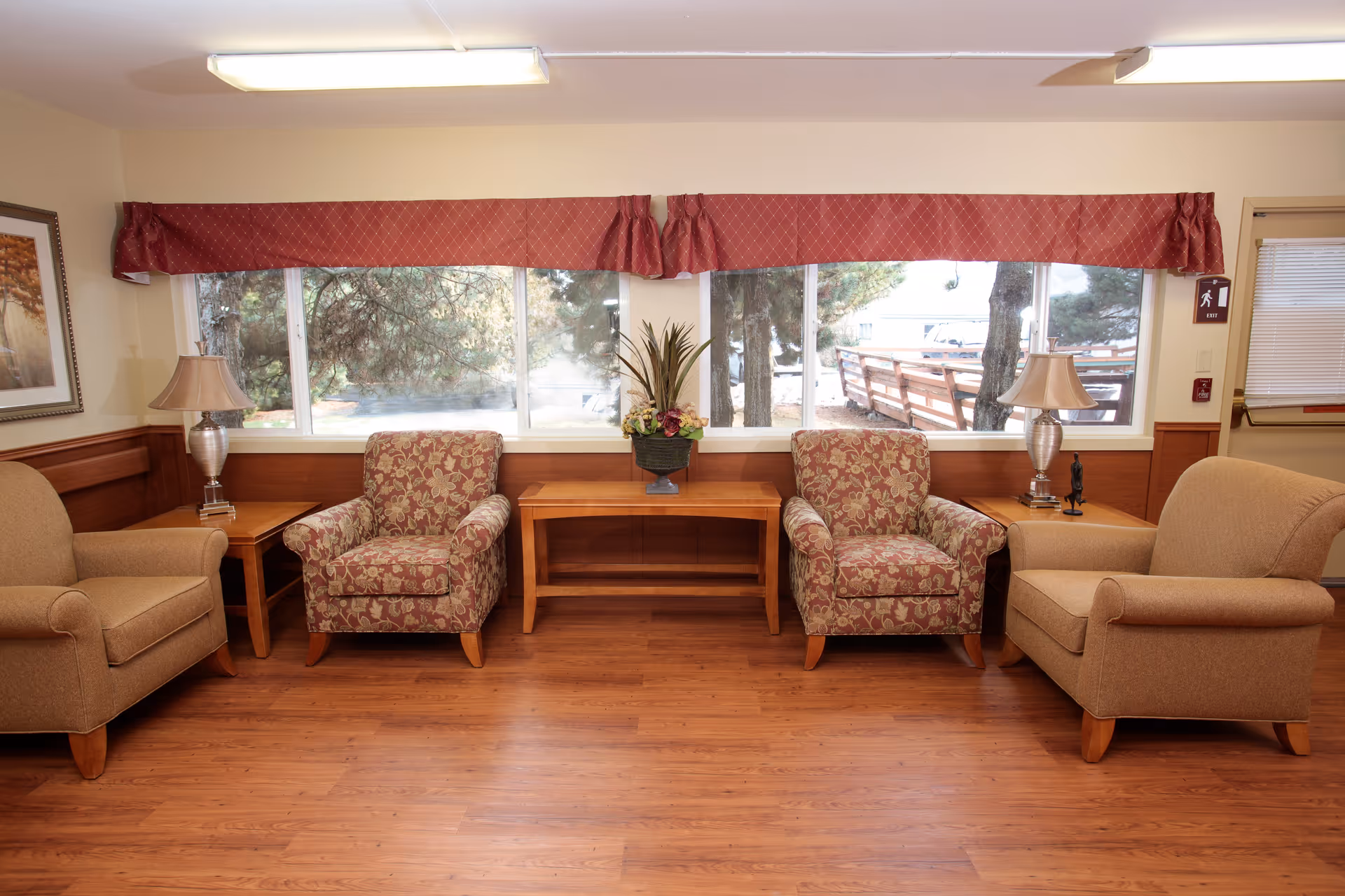 A cozy seating area with four armchairs arranged around two wooden side tables and a wooden coffee table with a flower arrangement. Large windows with red valances allow natural light to fill the room, showing trees and an outdoor walkway outside. The room has wooden flooring and beige walls with a framed picture on the left wall.
