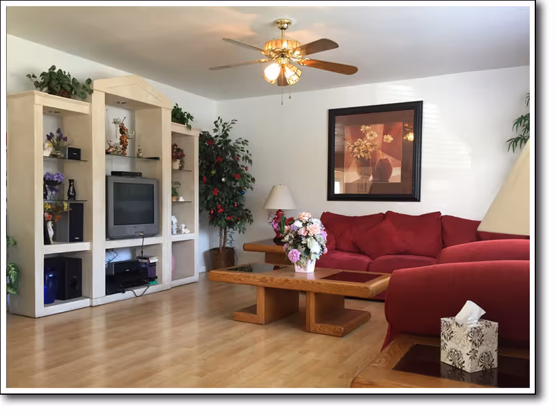 A living room with a red sofa and matching armchair, a wooden coffee table with a flower arrangement, a wooden entertainment center with a TV and decorative items, a ceiling fan with lights, a floor lamp, and a framed floral painting on the wall.