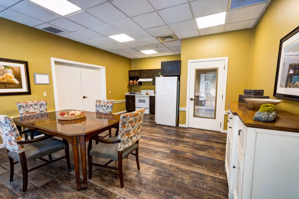Interior view of a dining area with a wooden table and six patterned chairs. The room has yellow walls, a white refrigerator, a stove, and dark wooden cabinets in the kitchen area. There is a white door with a glass panel and a white sideboard with decorative items on top.