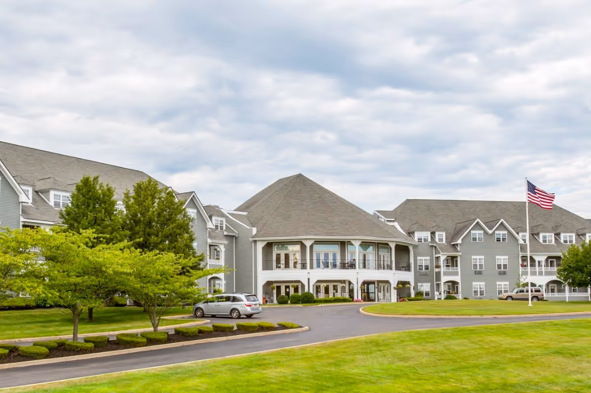 Exterior view of The Village at Mariner's Point senior living facility with a large gray building, green lawn, trees, a driveway with parked cars, and an American flag on a flagpole under a cloudy sky.