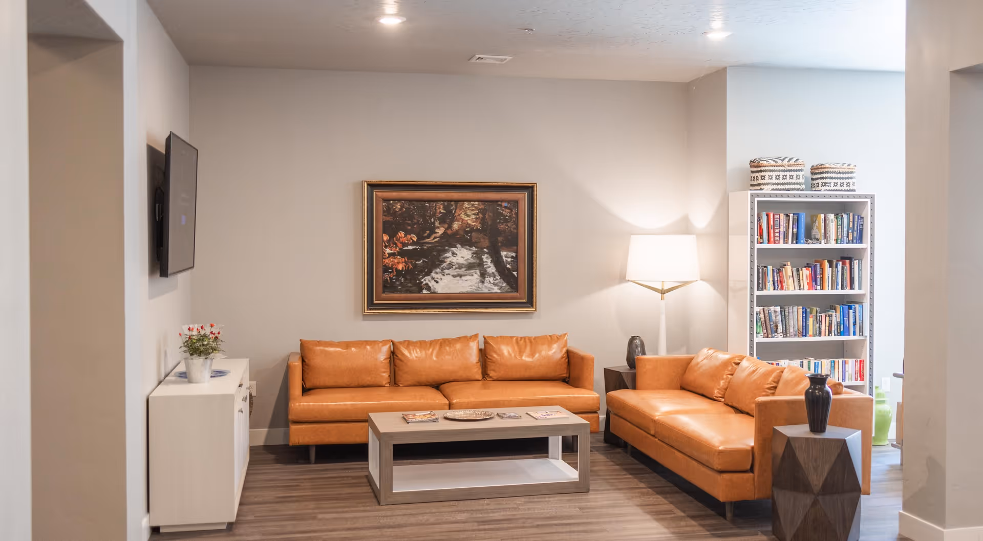 A cozy living room with two tan leather sofas arranged around a rectangular coffee table. A framed painting of a forest scene hangs on the wall above one sofa. To the right, there is a white bookshelf filled with books and two patterned baskets on top. A floor lamp with a white shade stands between the bookshelf and the sofa. A small side table with a black vase is next to the sofa on the right. The room has light-colored walls and wood flooring.