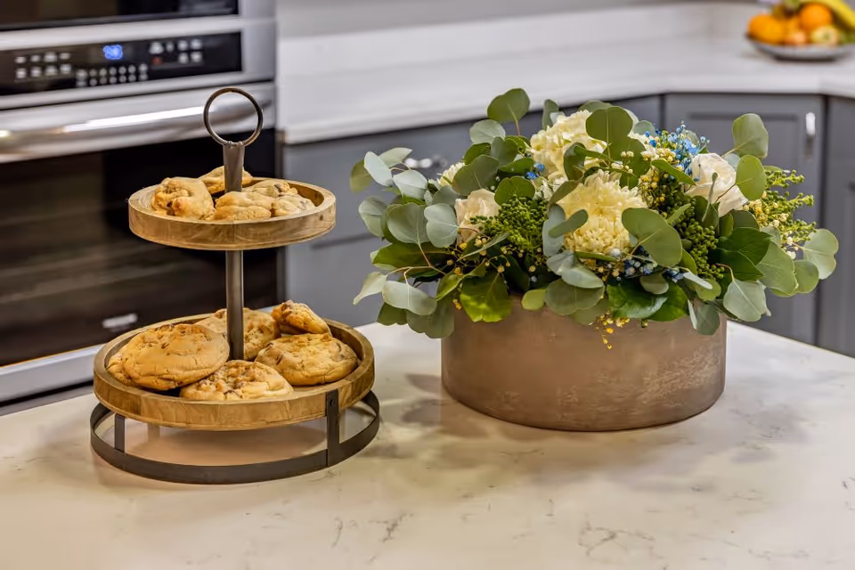 A kitchen countertop with a two-tier wooden stand holding chocolate chip cookies on the left and a round metal vase filled with a floral arrangement of white flowers and green leaves on the right. In the background, there is a stainless steel oven and gray kitchen cabinets.