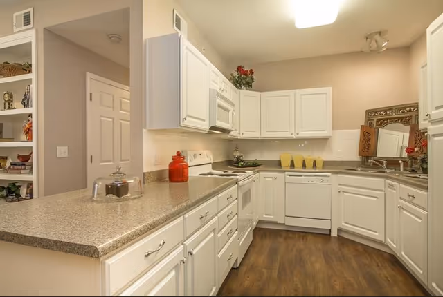 A clean, modern kitchen with white cabinets, a beige countertop, and wood flooring. The kitchen features a stove, microwave, dishwasher, and a sink with a decorative mirror above it. There are decorative items such as a red jar, a cake stand with a glass cover, and plants on the countertop and cabinets.