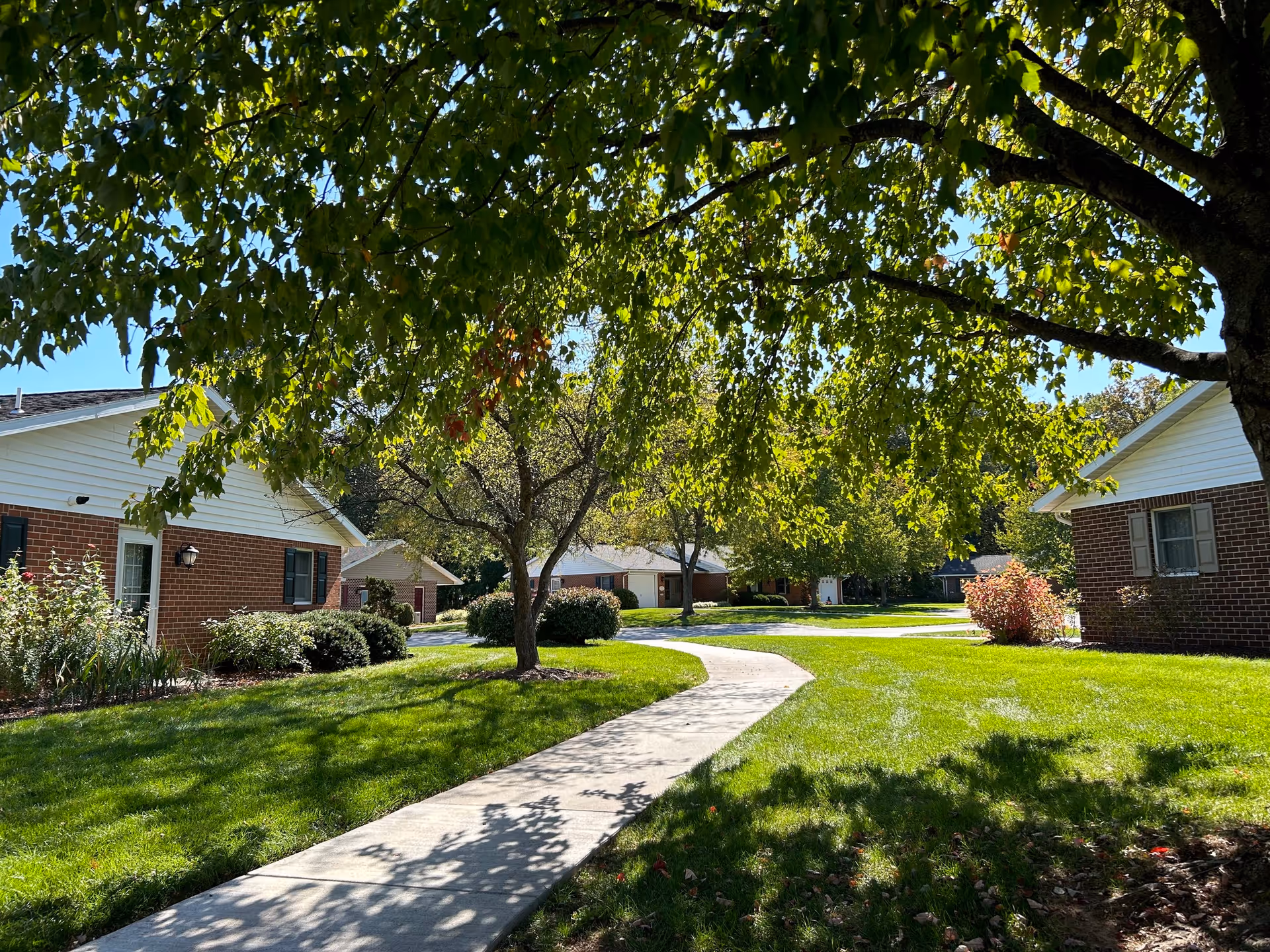 A sunny outdoor scene at Cross Keys Village featuring a curved concrete walkway surrounded by green grass, trees with green leaves casting shadows, and brick residential buildings with white siding and windows on either side.