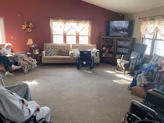 A cozy senior living common room with several elderly residents seated in chairs arranged around the perimeter. The room has a beige carpet, a large window with lace curtains, a TV on a wooden stand, and a maroon accent wall with a floral decoration.