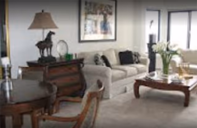 A cozy living room featuring a beige sofa with black and white cushions, a wooden coffee table with a vase of white flowers, a wooden side table with a lamp and horse figurine, a round wooden dining table with chairs, and large windows letting in natural light.