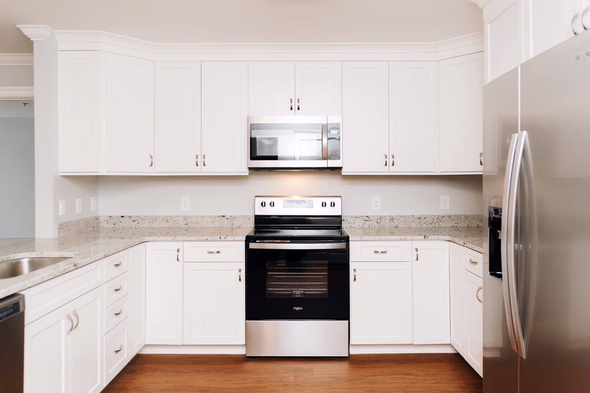 A modern kitchen with white cabinets, granite countertops, a stainless steel refrigerator, a stainless steel oven with a black glass door, and a stainless steel microwave above the oven. The kitchen has a wooden floor and a clean, bright appearance.