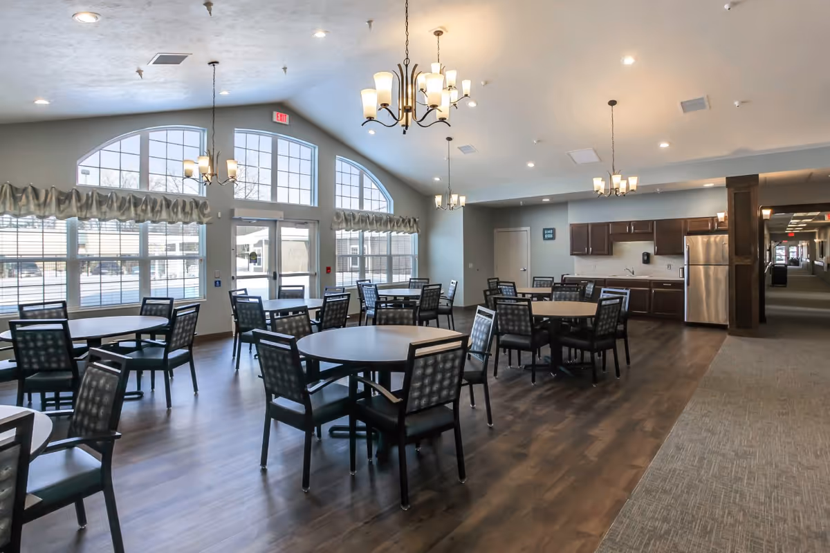 A spacious dining room with multiple round tables and chairs arranged neatly on a wooden floor. Large windows with arched tops allow natural light to fill the room. There are chandeliers hanging from the ceiling, and a kitchen area with dark wood cabinets and stainless steel appliances is visible in the background. A hallway extends to the right side of the room.