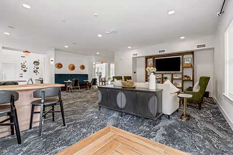 A spacious and modern common area in a senior living facility with a mix of seating options including bar stools, armchairs, and sofas. The room features a large built-in entertainment center with a TV, decorative shelves, and a console table with vases. The flooring is a combination of patterned carpet and wood-like tiles, and the walls are painted white with minimalistic decor. Large windows allow natural light to brighten the space.