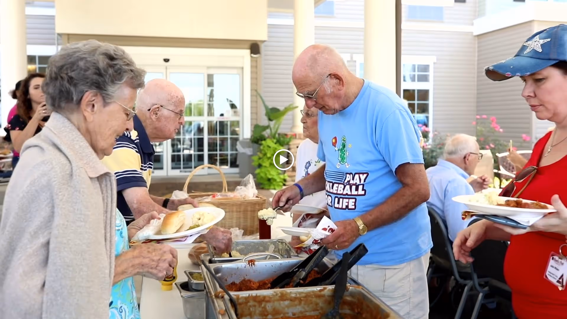 Several elderly people and a woman serving themselves food buffet-style outdoors at a senior living facility. The setting appears to be a patio area with tables and chairs, and the people are holding plates with food.