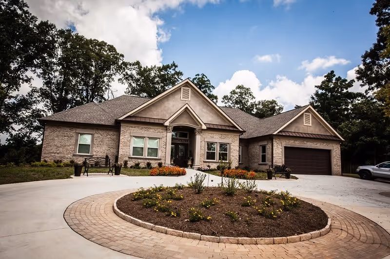 Front exterior view of a single-story brick residential building with a circular driveway and landscaped garden bed in the center. The building has a gabled roof, a covered entrance with columns, and a two-car garage. Trees and a partly cloudy sky are visible in the background.