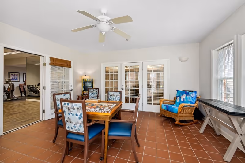 A bright room with terracotta tiled floor featuring a wooden table with a puzzle on it surrounded by four chairs with patterned cushions. There is a wicker rocking chair with a blue floral cushion near two windows on the right. Double glass doors with blinds lead to another room, and an open door on the left reveals exercise equipment in an adjacent room. A ceiling fan is mounted on the white ceiling.