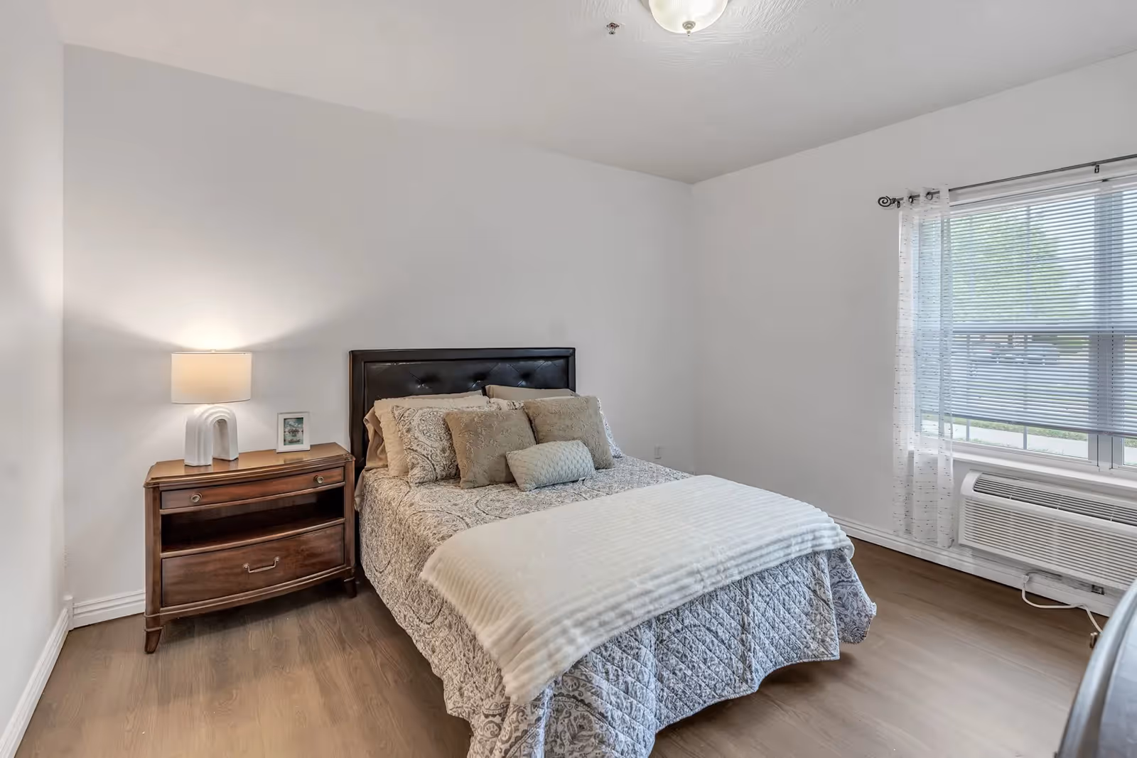 A neatly made bed with decorative pillows, a wooden nightstand and lamp sits in a bright, minimally furnished bedroom with a window and air conditioner.