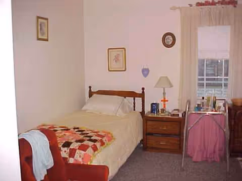 A furnished senior living bedroom featuring a bed, quilt, dresser, nightstand, and window.