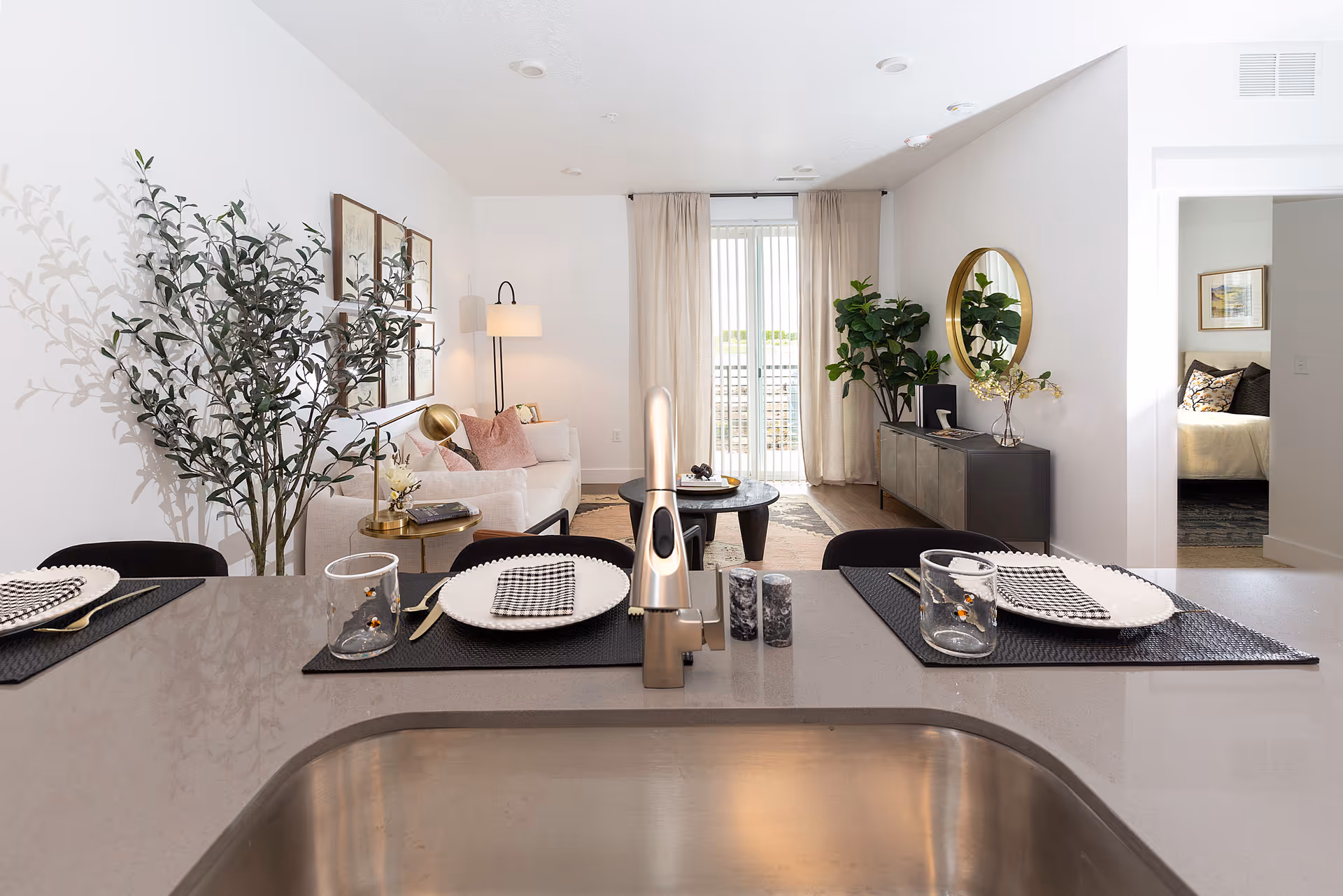 View from a kitchen island across a modern open-plan living room with place settings on the counter, a sofa, plants, and a console with a round mirror.
