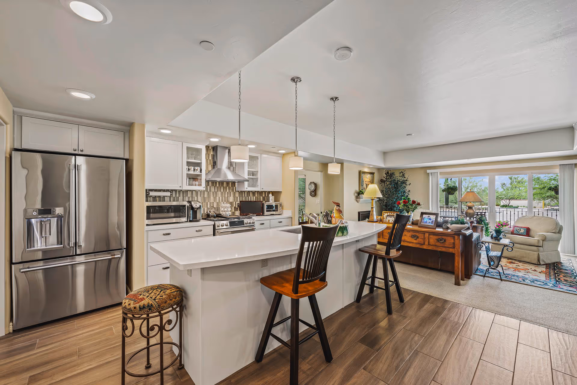 Open kitchen and living room area with a large white island counter featuring two wooden bar stools. The kitchen has white cabinets, stainless steel refrigerator, stove, microwave, and pendant lights hanging from the ceiling. The living room has a wooden console table with framed photos, a lamp, an armchair, and large sliding glass doors leading to an outdoor patio.