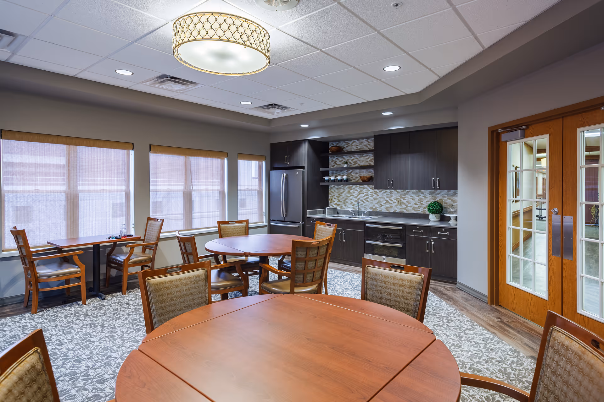 A well-lit dining area with round and rectangular wooden tables surrounded by chairs. The room features large windows with blinds, a modern kitchen area with dark cabinets, a stainless steel refrigerator, oven, and a decorative backsplash. There are double wooden doors with glass panels on the right side.
