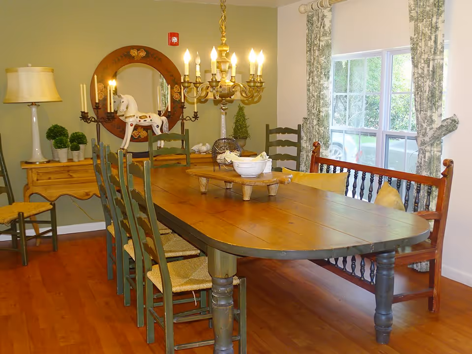 A cozy dining room with a large wooden dining table surrounded by green wooden chairs and a wooden bench with yellow cushions. The room features a chandelier with lit candles hanging above the table, a round mirror with floral designs on the wall, a side table with a lamp, potted plants, and decorative items including a white horse figurine. A window with floral curtains lets in natural light.