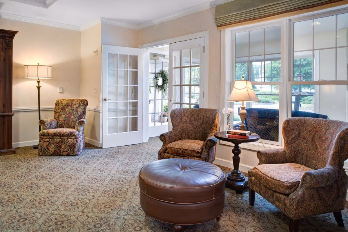 Bright sitting room with patterned armchairs, a round leather ottoman, a side table with a lamp, and French doors leading outside.