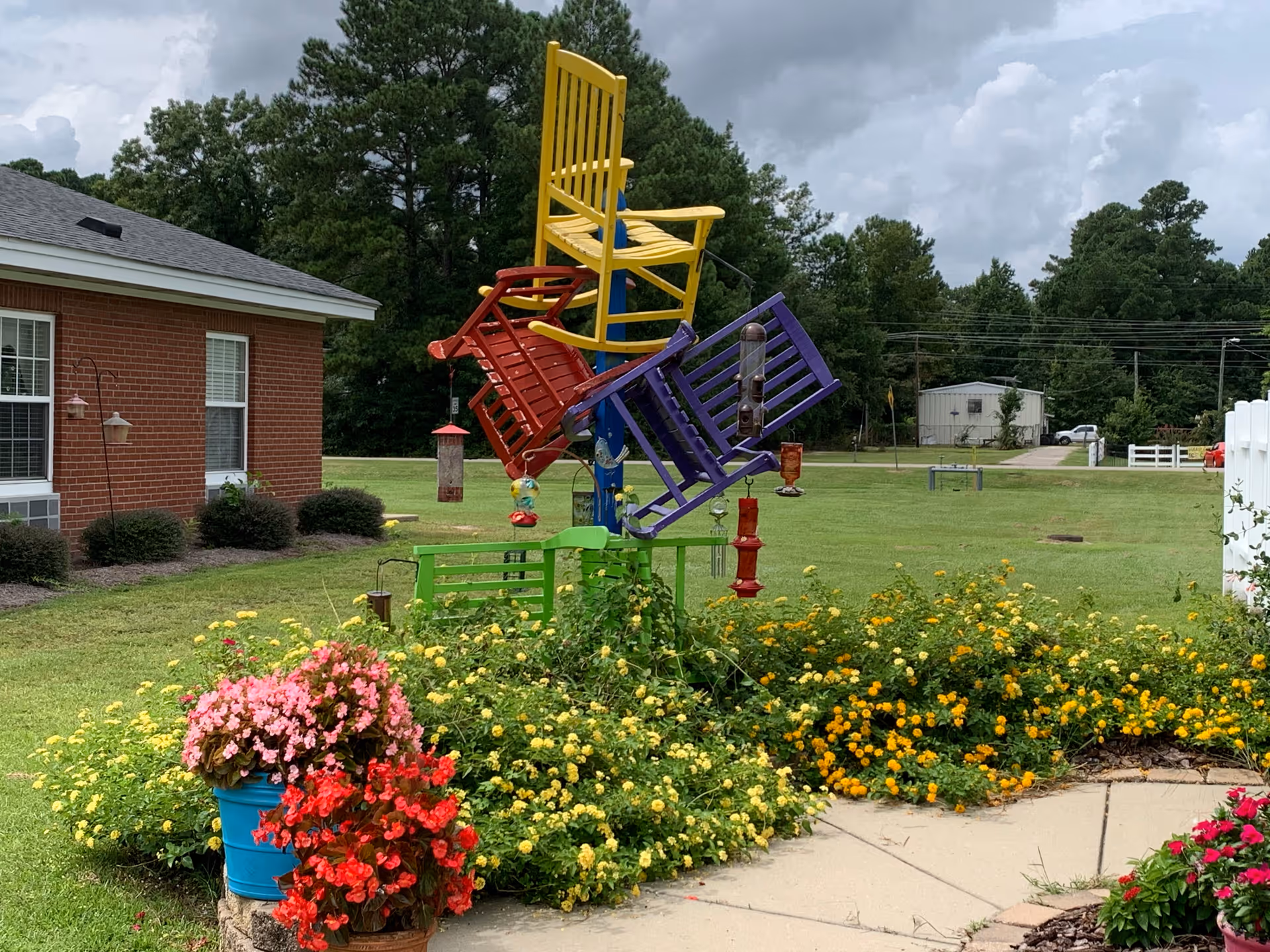 Outdoor garden area at Open Arms Retirement Center featuring a colorful birdhouse structure made from painted wooden chairs in yellow, red, purple, and green. The garden is filled with blooming flowers in red, pink, and yellow, with a brick building and green lawn in the background under a cloudy sky.