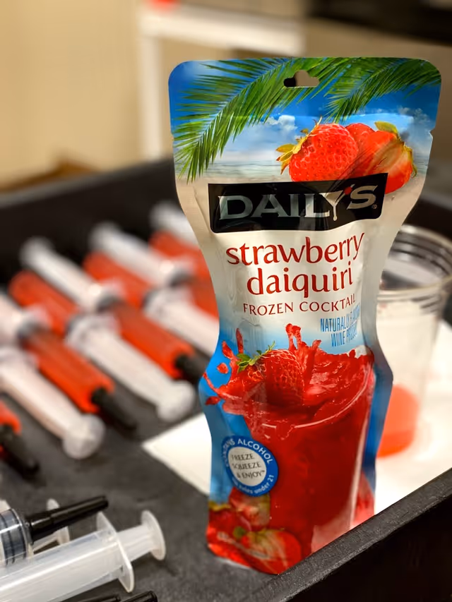 A pouch of Daily's strawberry daiquiri frozen cocktail standing on a tray alongside several syringes filled with red liquid.