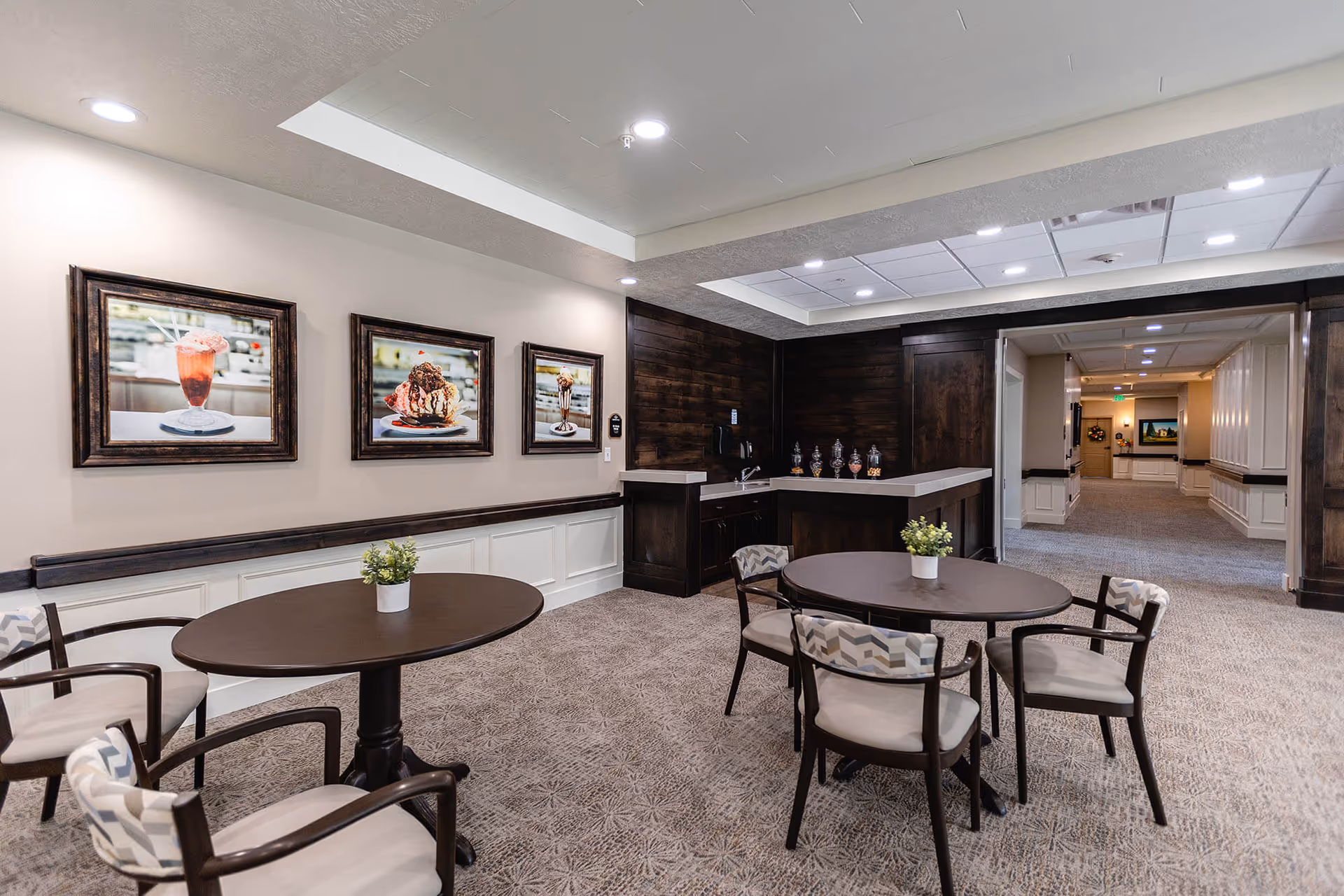 A cozy dining area in a senior living facility with two round dark wood tables, each surrounded by four cushioned chairs with patterned backs. Small potted plants are placed on each table. The walls are decorated with three framed pictures of desserts. There is a small counter area with a sink and jars of snacks in the corner. The space is well-lit with recessed ceiling lights and has carpeted flooring.