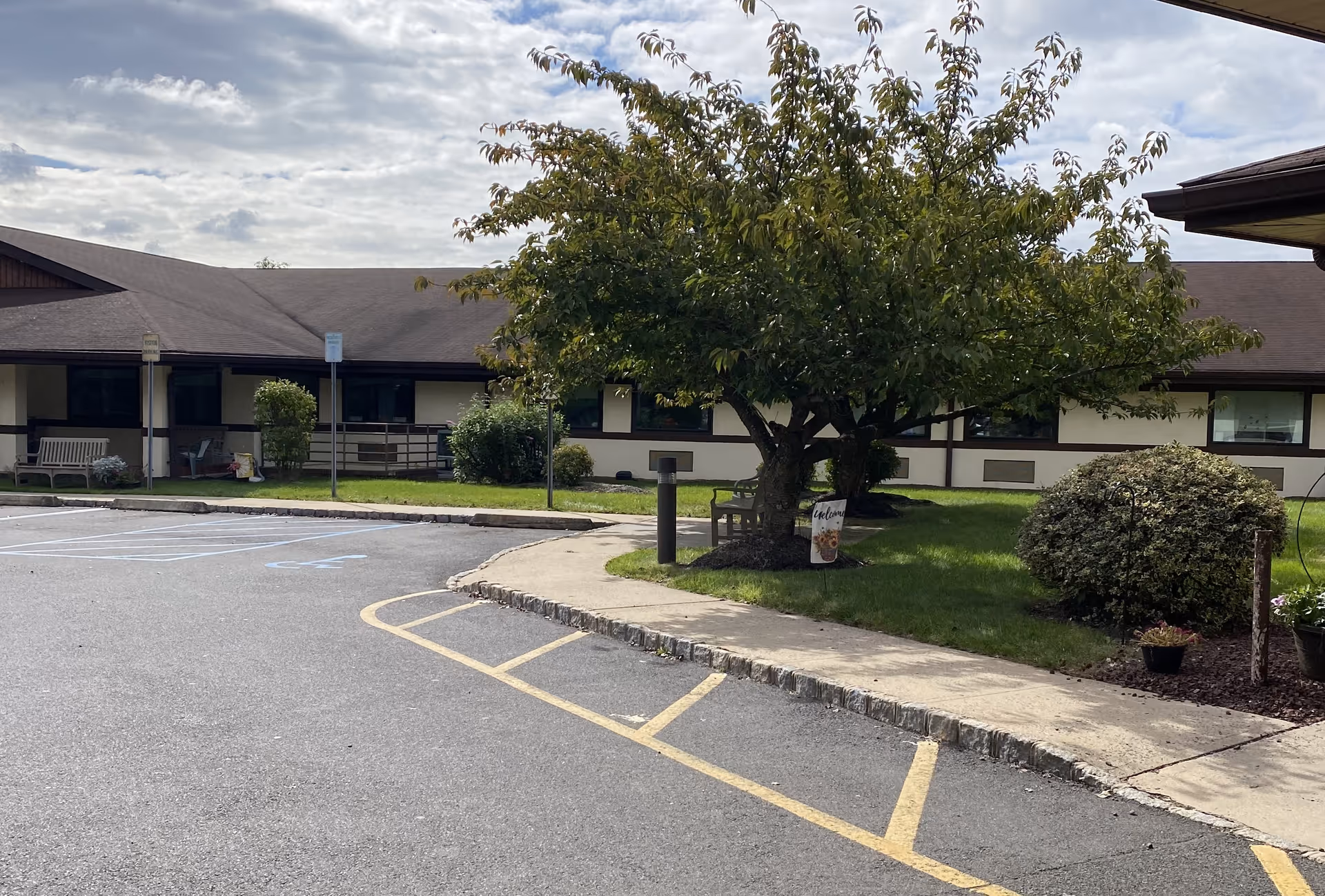 Exterior view of a single-story building with a brown roof and beige walls, surrounded by a parking lot with marked spaces and a sidewalk. There is a tree with green leaves and some bushes on a grassy area near the sidewalk, along with a bench and a small welcome sign. The sky is partly cloudy.