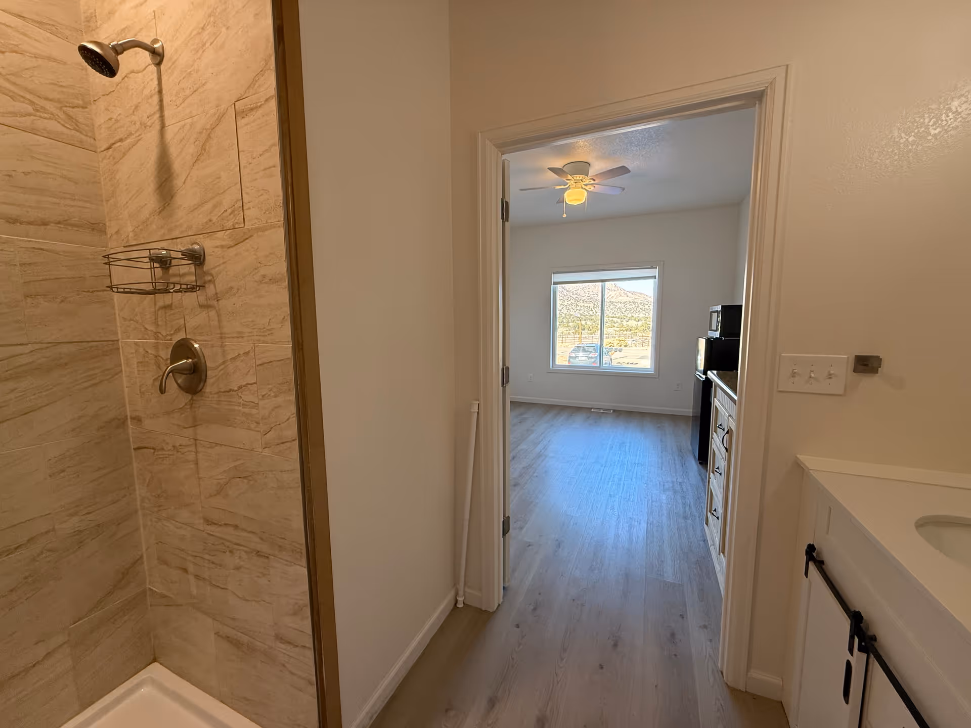 View from a bathroom showing a tiled shower with a showerhead and metal basket, a white vanity with a sink, and an open doorway leading to a room with wood flooring, a ceiling fan with light, a window with a view of a parked car and hills outside, and a small kitchenette area with a microwave and mini fridge.