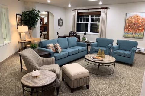 A cozy living room with a blue sofa, two matching blue armchairs, a striped armchair, and a striped ottoman. There are two round wooden coffee tables with metal legs, one holding a tissue box and the other with decorative items. A window with beige curtains and a colorful painting of trees adorn the wall. A lamp on a side table and a potted plant add warmth to the space.