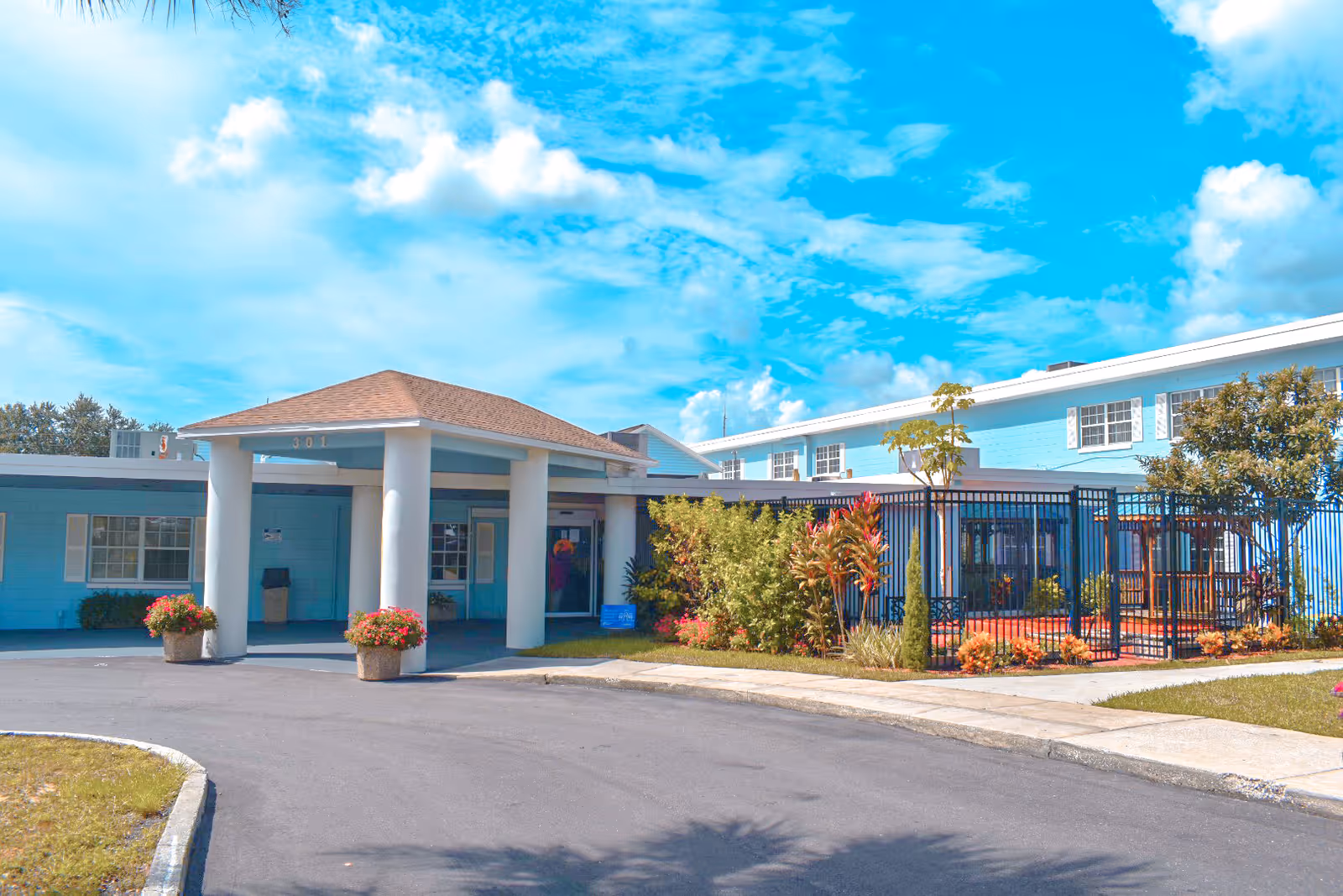 Exterior view of a light blue assisted living facility building with a covered entrance supported by four white columns. There are potted plants with flowers near the entrance and a fenced garden area with various shrubs and small trees. The sky is bright blue with scattered clouds.