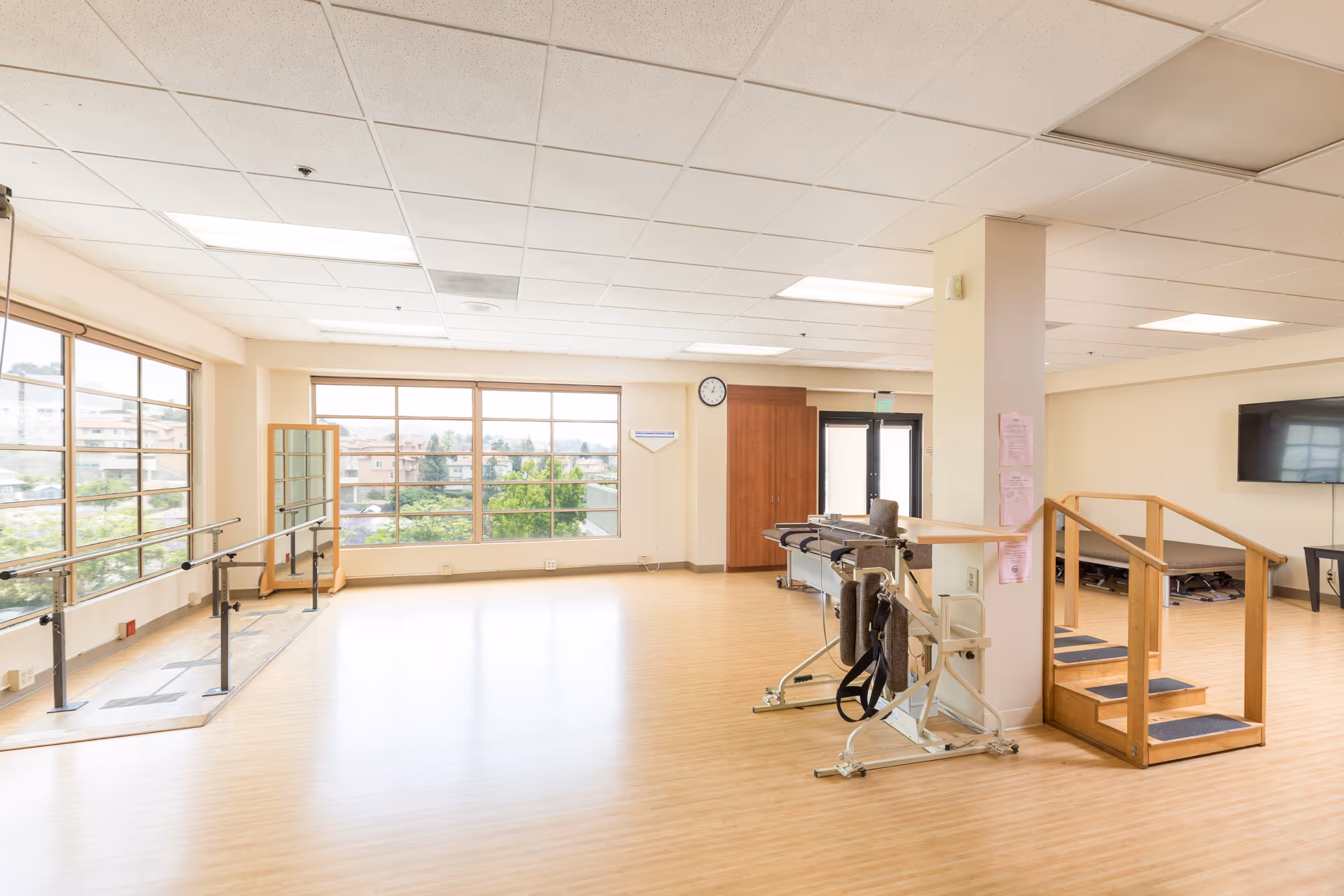 A spacious physical therapy room with large windows letting in natural light. The room features parallel bars for walking exercises, a wooden staircase with handrails for step training, padded therapy tables, and a wall-mounted TV. The floor is light wood, and the walls are painted a soft beige.