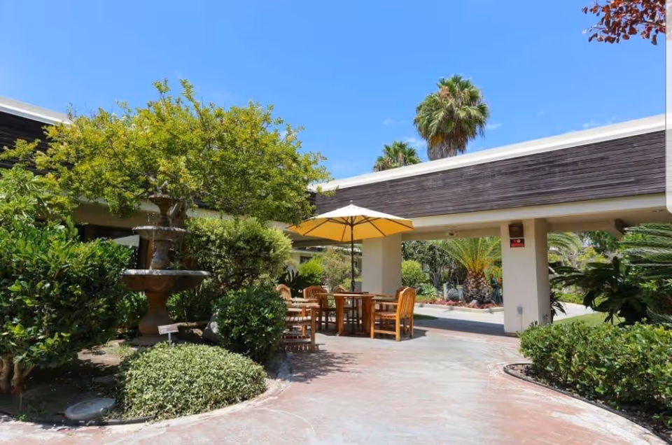 Outdoor patio area at Vista Knoll Specialized Care with wooden tables and chairs under a yellow umbrella, surrounded by green bushes and trees, including palm trees, under a clear blue sky.