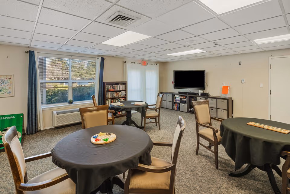 A well-lit common room with several round tables covered with black tablecloths, each surrounded by chairs. One table has a Chinese checkers game set up. There is a large window with blue curtains, a bookshelf filled with books, a flat-screen TV mounted on the wall, and storage cabinets beneath it. The room has a carpeted floor and a drop ceiling with fluorescent lighting.