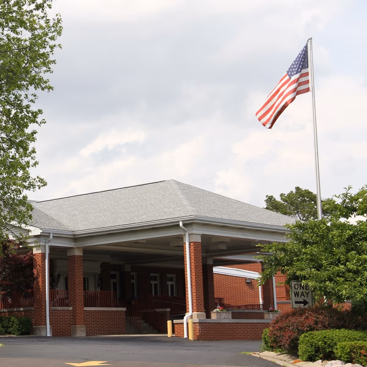 Exterior view of a brick building with a covered entrance supported by columns, an American flag on a tall flagpole, and surrounding greenery including trees and bushes under a cloudy sky.