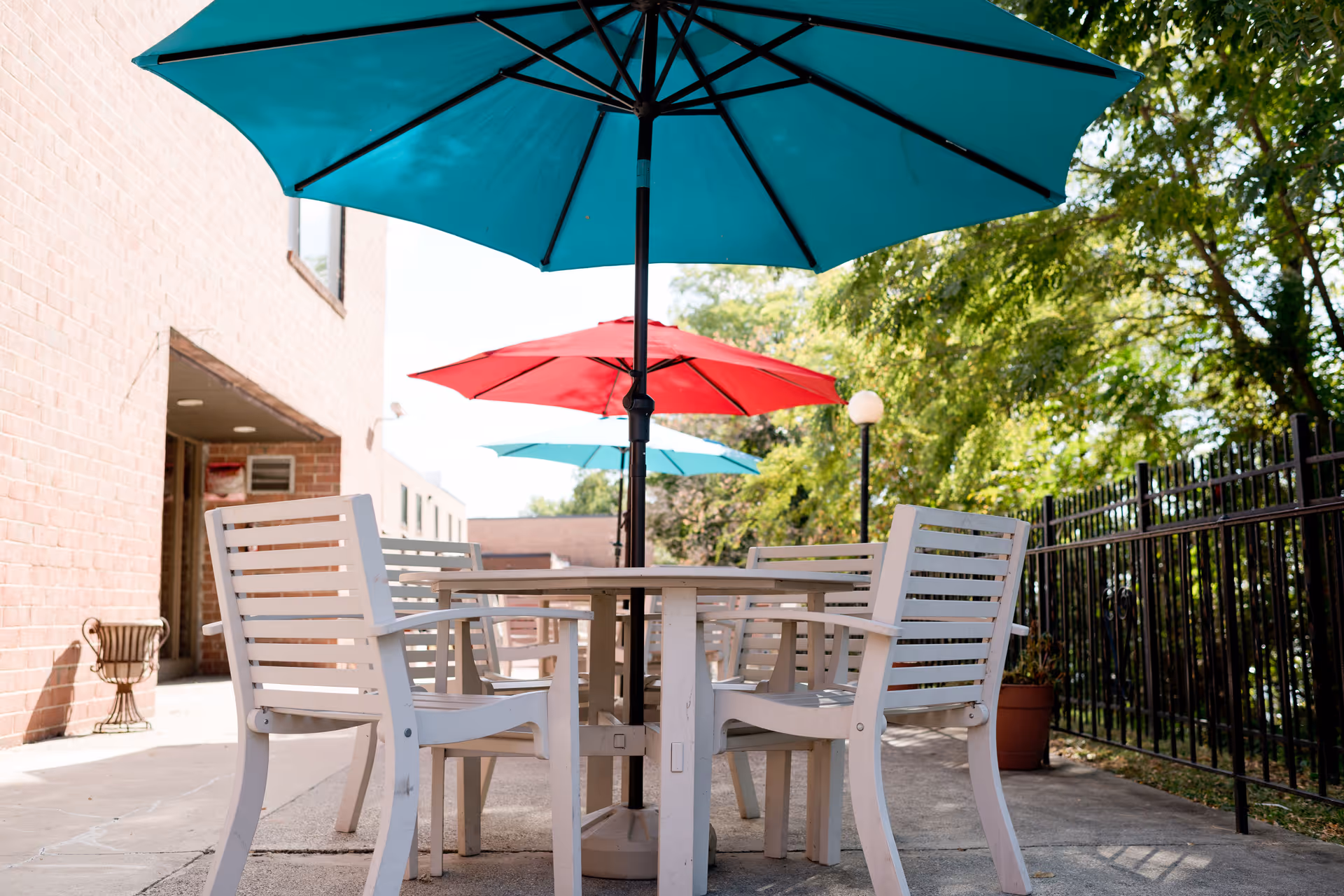 Outdoor patio area with white wooden chairs and tables shaded by large blue and red umbrellas, next to a brick building and surrounded by trees and a black metal fence.