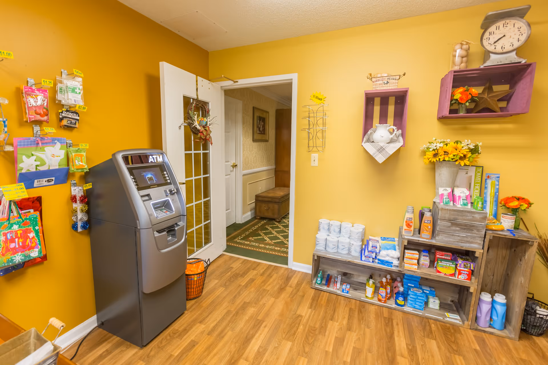 Interior view of a small convenience area with yellow walls and wood flooring. There is an ATM machine on the left side next to a wall with hanging small items for sale. On the right side, wooden crates hold various personal care products, paper towels, and flowers. A door with glass panels leads to a hallway with carpet and framed artwork.