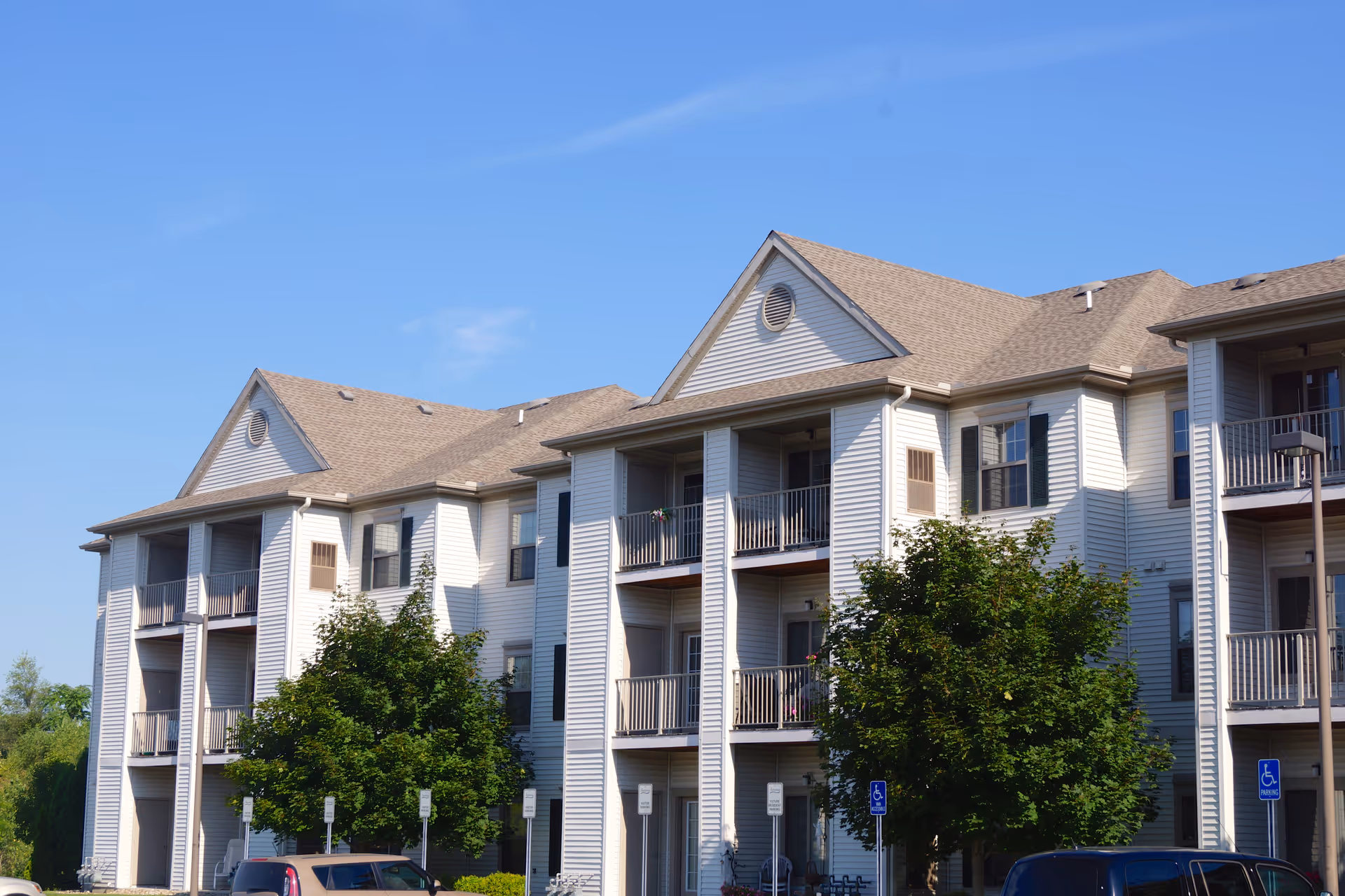 Exterior front of a three-story apartment building with balconies, trees, and parked cars under a clear blue sky.