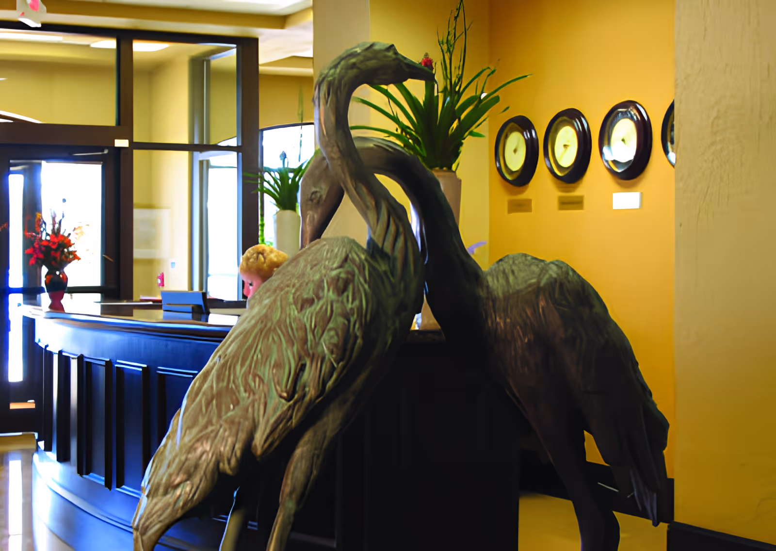 Interior view of a facility reception area with a dark wooden curved front desk. In front of the desk are two large decorative bird sculptures. Behind the desk, there are plants in vases and a wall with four round clocks showing different times. The walls are painted yellow and the area is well-lit with natural light coming through glass doors and windows.