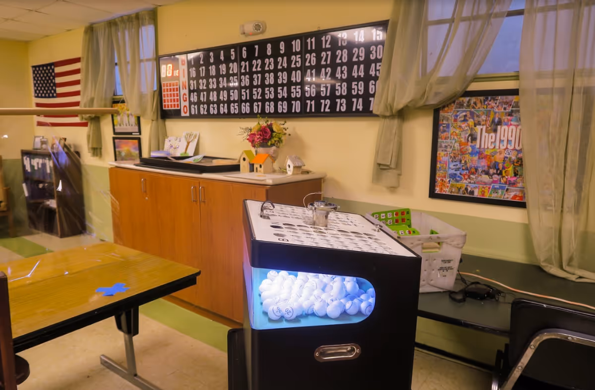 A senior activity room with a bingo machine filled with numbered balls, a large bingo board on the wall, tables, and decorations.