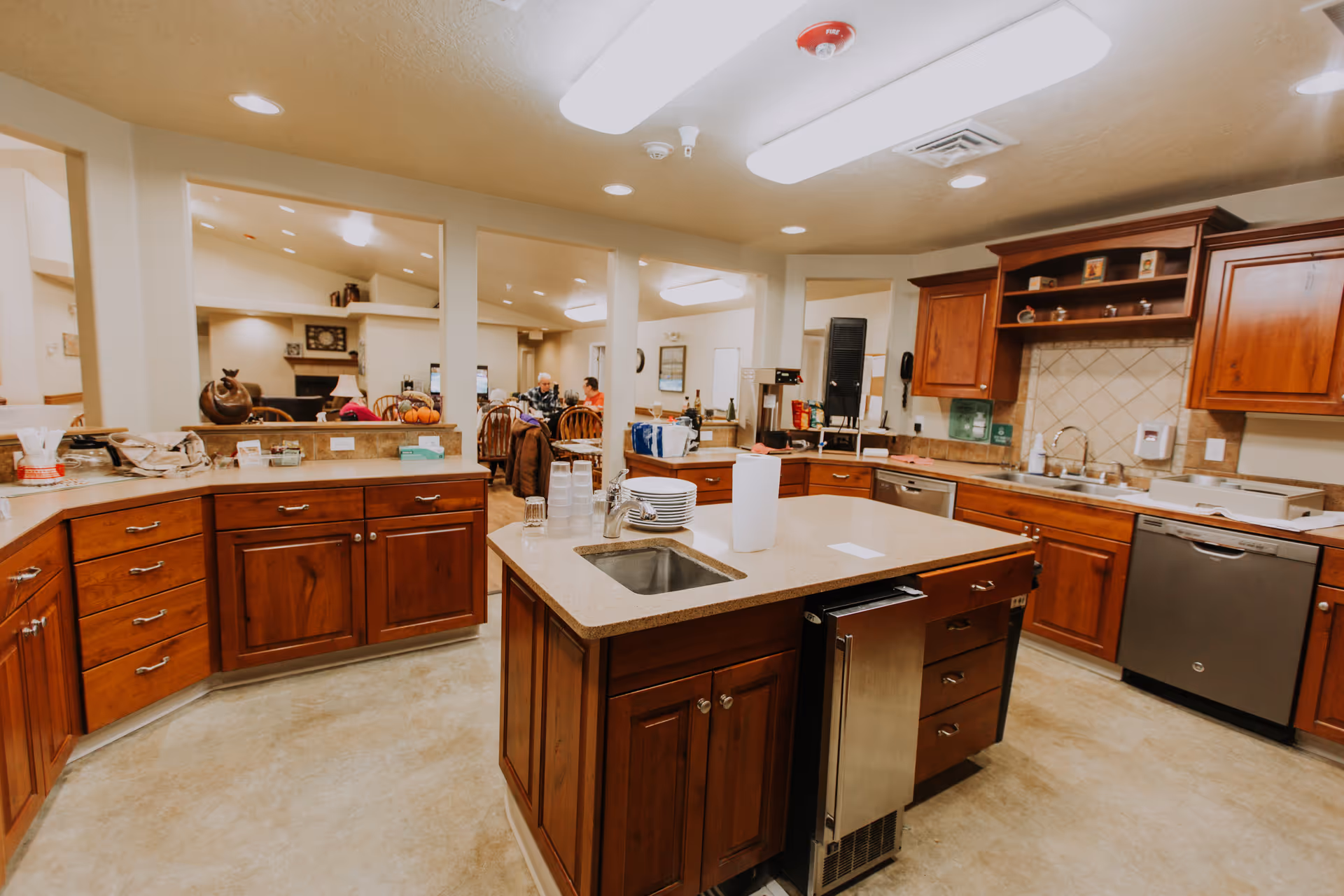 Open communal kitchen with wooden cabinets, a central island sink, and stainless steel appliances overlooking a dining area.