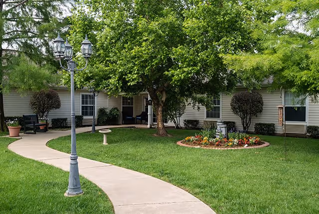 A well-maintained outdoor garden area at Morada Southridge featuring a curved concrete walkway, a vintage-style street lamp, a birdbath, a flower bed with colorful flowers, green grass, and a large leafy tree in front of a single-story building with beige siding and several windows.