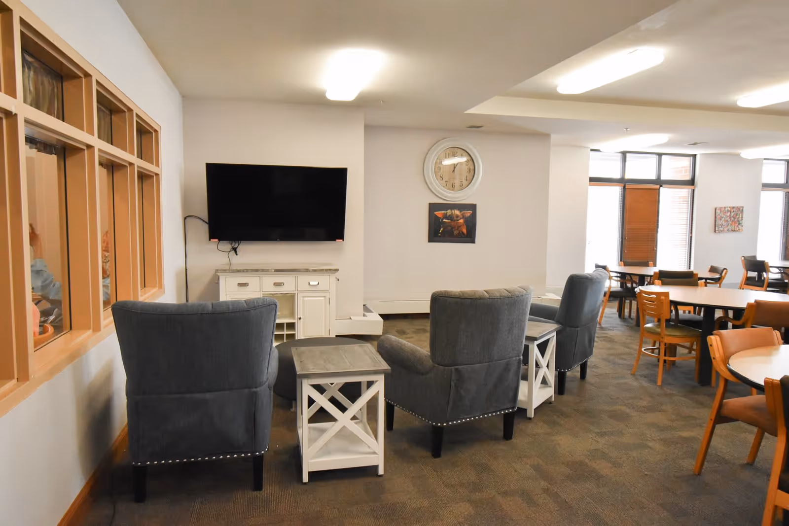 A senior living facility common area with three gray armchairs arranged around two small white side tables. A flat-screen TV is mounted on the wall above a white cabinet. A clock and a framed picture hang on the wall. In the background, there are several wooden tables and chairs near large windows with blinds.