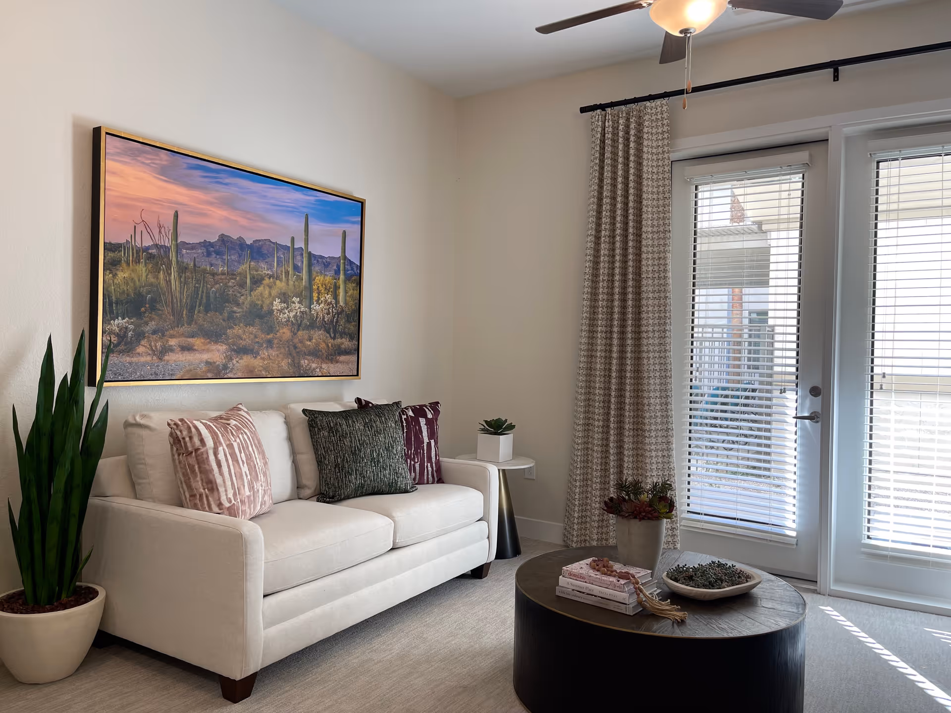A cozy living room with a white sofa adorned with three decorative pillows in different patterns and colors. Above the sofa hangs a large framed picture of a desert landscape with cacti and mountains during sunset. To the left of the sofa is a tall green potted plant. In front of the sofa is a round dark wooden coffee table with books and small potted plants on it. To the right, there are glass doors with blinds and a patterned curtain, letting in natural light.